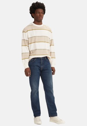 Chemise à rayures à manches longues dans les tons crème, beige et vert. Associée à un jean en denim bleu. Chaussures blanches en baskets. Design simple, coupe décontractée.