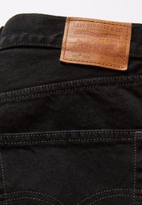 Pantalones vaqueros negros de mezclilla con una etiqueta de cuero marrón con detalles en relieve. Se pueden ver puntadas distintivas y diseño de bolsillos.
