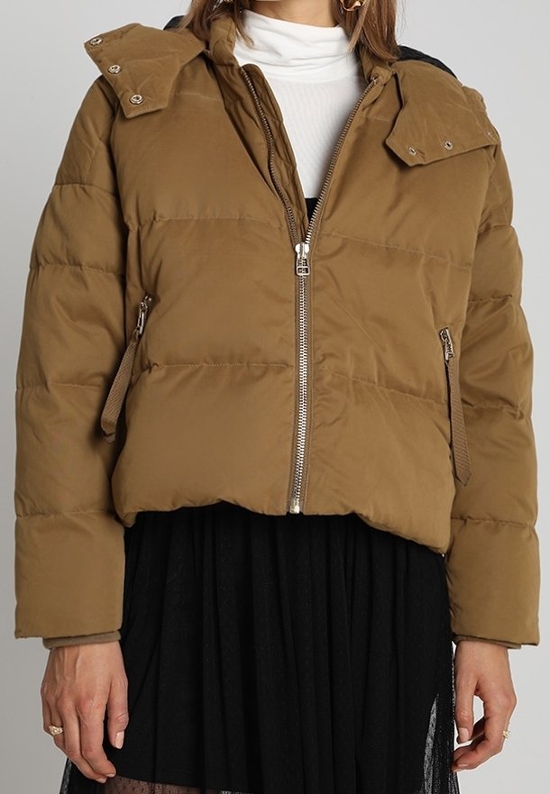 Femme portant une veste matelassée marron avec fermeture éclair frontale, col à boutons-pression et poches latérales zippées, par-dessus un col roulé blanc et une jupe noire.