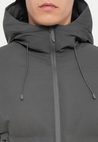 Detailný záber na osobu v tmavošedej mikine s kapucňou, ktorá má predný zips a z kapucne visia šnúrky.