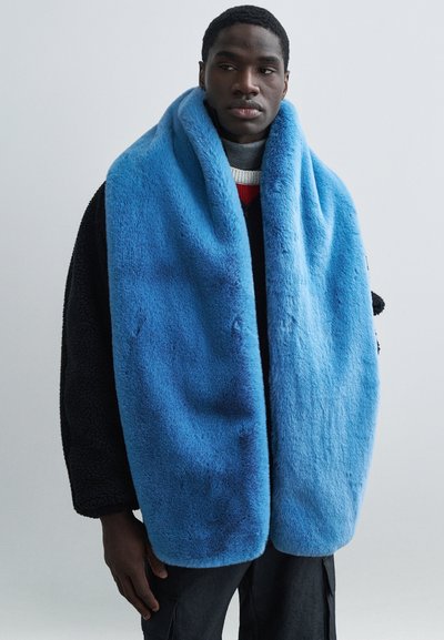 Μπλε κασκόλ από ψεύτικη γούνα με απαλή υφή, το οποίο έχει τυλιχτεί γύρω από το λαιμό, συνοδευόμενο από ένα σκούρο στρωματοποιημένο εξωτερικό ένδυμα και ένα πουλόβερ με κόκκινες λεπτομέρειες.