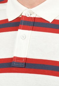 Poloshirt in het wit met rode en marineblauwe horizontale strepen. Beschikt over een klassieke kraag en twee knopen. Zachte, lichte stof met een gemiddelde textuur.