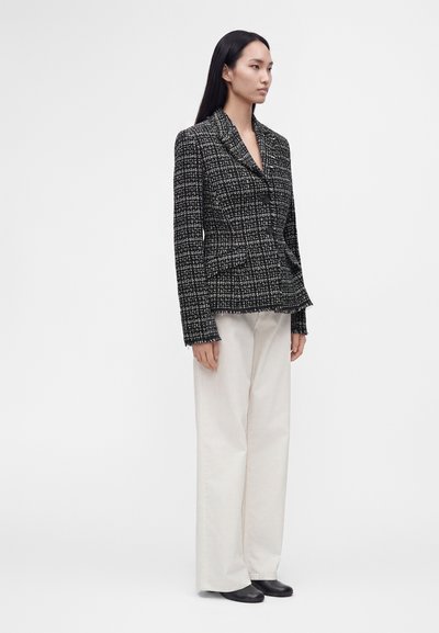 Femme portant un blazer texturé noir et blanc, un pantalon blanc à jambes larges et des chaussures plates noires, debout devant un fond clair uni.