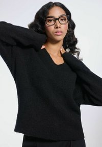 Czarny, luźny sweter z miękkiego, teksturowanego materiału z dekoltem w kształcie litery V i długimi rękawami. Model pozuje z uniesionymi rękami na jednolitym tle.
