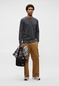 Szary sweter z dzianiny, beżowe spodnie cargo, koszula w kratę zarzucona na ramię, czarna torba na ramię, srebrne buty sportowe. Prosty, casualowy strój.