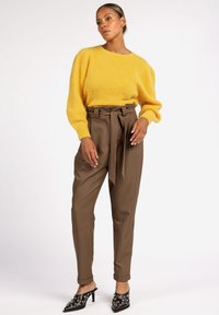 Vrouw die staat in een gele fluffy trui, hoge bruine paperbagbroek met riem, en zwarte gespijkerde puntige hakken.