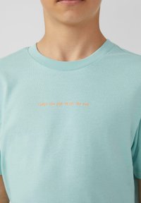 Lichtblauwe katoenen t-shirt met een ronde hals en korte mouwen. De voorkant toont oranje tekst: "Ververs de zon en geniet van het plezier."