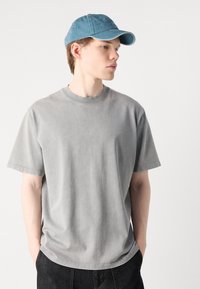 Gray cotton t-shirt with short sleeves, round neckline, and a relaxed fit. Worn with a light blue denim cap. Plain design, no patterns.
