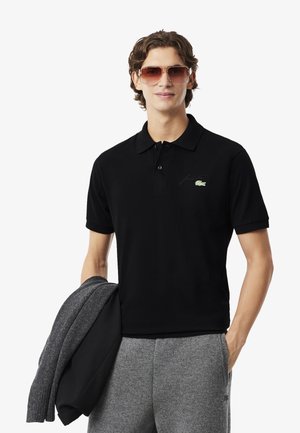 Polo noir avec un col côtelé, des manches courtes et un logo brodé vert. Le modèle tient un pull en maille gris. Lunettes de soleil marron.
