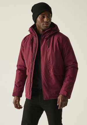 Mężczyzna w bordowej kurtce z kapturem, czarnej czapce, czarnej koszuli i czarnych spodniach, stojący z rękami swobodnie opuszczonymi wzdłuż boków.