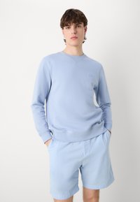 Felpa azzurra in materiale morbido con maniche lunghe, scollatura a girocollo e una sottile toppa con logo sul petto, abbinata a pantaloni corti coordinati.
