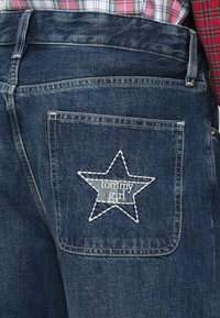 Jeans in denim blu scuro con una stella ricamata bianca sulla tasca posteriore. La tasca presenta il testo "tommy girl" all'interno del design della stella.