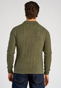 Groene kabelgebreide trui met een hoge halslijn, geribbelde manchetten en zoom, met een gestructureerd patroon. Het model draagt donkere jeans.