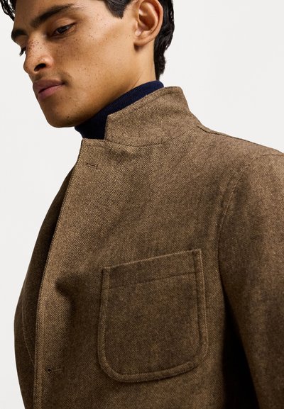 Brauner Fischgrätmantel mit hohem Kragen und einer Brusttasche, aus strukturiertem Stoff gefertigt. Mit einer figurbetonten Passform und einem navyblauen Rollkragenpullover darunter.