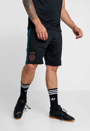 Männlich, trägt schwarze Ajax-Sportshorts mit türkisfarbenen Streifen, schwarze Adidas-Socken und schwarze Hallenfußballschuhe, steht vor einem hellen Hintergrund.