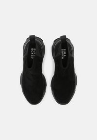 Svarta skopresenter i mocka med en slät struktur, som har en tjock gummisula och varumärkeslogo på insidan av tungan.