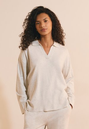 Donna con capelli ricci che indossa un maglione beige chiaro a maniche lunghe con colletto e pantaloni abbinati, in piedi con le mani in tasca su uno sfondo semplice.