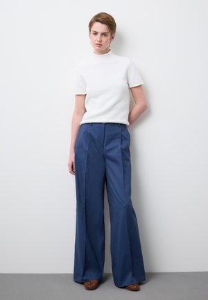 Mujer de pie junto a una pared, vestida con una blusa blanca de manga corta, pantalones anchos azul de tiro alto, y zapatos marrones trenzados.