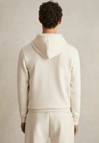 Hoodie u krem boji napravljen od mekog materijala, s kapuljačom na vezanje i rebrastim manžetama. Glatka tekstura i opušteni kroj.