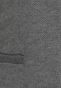 Detailní záběr na šedou tkaninu s jemným vzorem rybí kosti a malým textilním poutkem.