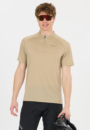 Junger Mann trägt beiges kurzärmeliges Fahrradtrikot und schwarze Shorts, hält einen schwarzen Fahrradhelm und trägt reflektierende Sportsonnenbrille.