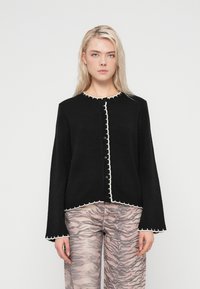 Μαύρο πουλόβερ με λευκή διακοσμητική κυματοειδή λεπτομέρεια, στρογγυλή λαιμόκοψη και κουμπιά ως κλείσιμο. Μακριά μανίκια με φαρδιά άκρα. Φορεμένο με ανοιχτόχρωμο παντελόνι με σχέδια.