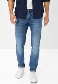 Jeans di denim azzurro chiaro con taglio dritto, cuciture visibili e risvolti. Abbinati a sneakers bianche e una camicia blu navy.