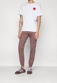 Wit katoenen T-shirt met rode cirkelgrafiek op de linkerborst, gecombineerd met lichtbruine cargobroek met zijzakken en taps toelopende pijpen. Bruine sneakers.