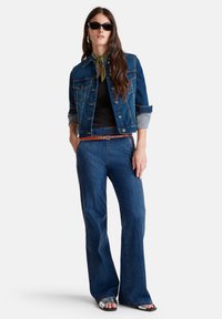 Femme portant une veste en jean foncé et un jean, un haut noir, un foulard vert au cou, des lunettes de soleil, une ceinture marron et des sandales métalliques, se tenant avec les mains dans les poches.