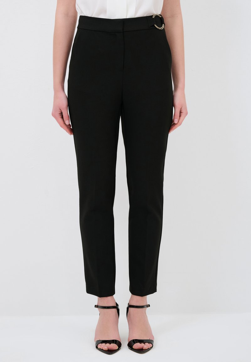 Zwarte op maat gemaakte broek met een strak ontwerp, voorzien van een ronde gouden hardwareaccent op de taille en taps toelopende broekspijpen. GLadde stoffen structuur.
