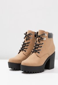 Botines beige con parte superior sintética, cordones negros, ojales plateados y un tacón grueso. Suela negra texturizada con patrones de relieve.