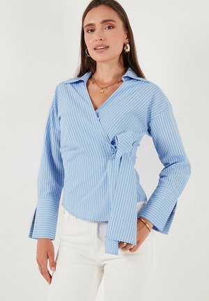 Woman wearing a light blue and white striped wrap blouse with a bow tie on the side, paired with white pants and gold jewelry.
