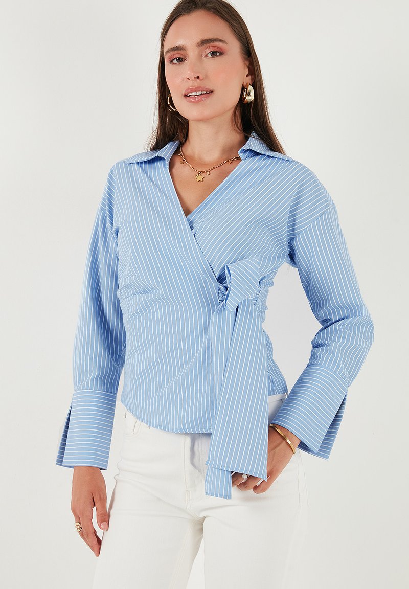Donna che indossa una blusa avvolgente a righe blu chiaro e bianche con un fiocco laterale, abbinata a pantaloni bianchi e gioielli dorati.