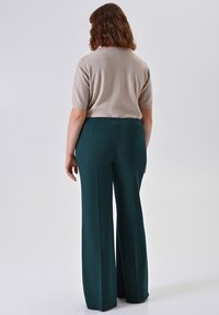 Vrouw met schouderlang golvend haar, gekleed in een beige korte mouwen top en donkergroene wijde broeken, staand tegen een effen achtergrond.