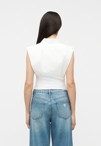 Γυναίκα με μακριά σκούρα μαλλιά, που φοράει αμάνικη λευκή μπλούζα  με δομημένους ώμους και μπλε ψηλόμεσα, φαρδιά τζιν, κοιτάζοντας προς τα πίσω.