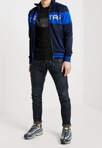 Chaqueta azul marino con cremallera y franjas azul claro, camiseta negra con gráfico, vaqueros oscuros y zapatillas grises con detalles amarillos.