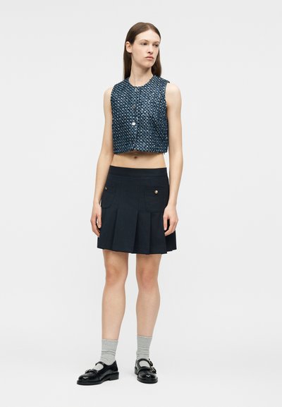 Νεαρή γυναίκα με μπλε αμάνικο κοντό υφασμάτινο γιλέκο, πλισέ σκούρο μπλε φούστα με τσέπες, γκρι κάλτσες και μαύρα παπούτσια Mary Jane, στέκεται ακίνητη.