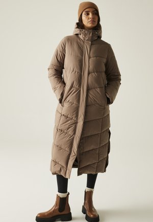 Kobieta ubrana w długi brązowy pikowany płaszcz z kapturem, brązową dzierganą czapkę, czarne legginsy i brązowe botki, stojąca z rękami w kieszeniach.