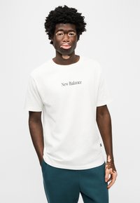 Λευκό βαμβακερό t-shirt με γκρι λογότυπο κειμένου "New Balance", κοντά μανίκια και άνετη εφαρμογή, συνδυασμένο με σμαραγδί φόρμα.