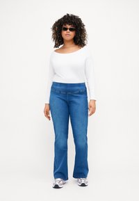 Vaqueros acampanados azules con una textura suave, que cuentan con una cinturilla discreta y costuras en los bolsillos frontales. Combinados con una blusa blanca de manga larga.