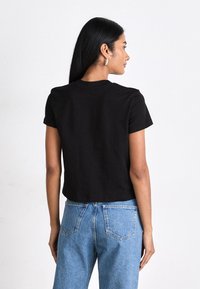 Camiseta negra cortada con mangas cortas, hecha de algodón suave. La parte posterior presenta un escote sin costuras y un diseño sencillo. Se combina con unos vaqueros de mezclilla azules.