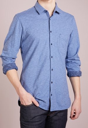 Mann trägt ein langärmliges blaues Hemd mit aufgekrempelten Ärmeln und dunkle Jeans, steht mit einer Hand in der Tasche.