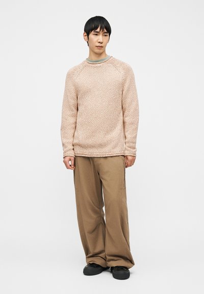 Homme portant un pull texturé beige clair, un pantalon marron ample et des chaussures noires, debout devant un fond blanc uni.