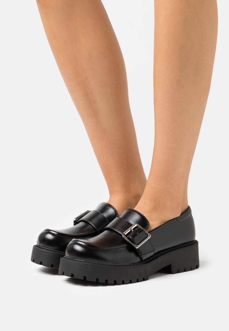 Jambes portant des mocassins à plateforme épaisse noirs avec un grand détail de boucle en argent et des semelles épaisses texturées sur un fond clair.