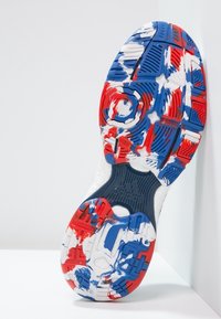 Suola della scarpa con un design testurizzato in rosso, bianco e blu, con scanalature decorative e un elemento centrale di presa circolare.