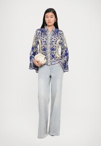 Bluză cu model floral în culori alb și albastru, cu mâneci evazate, împerecheată cu blugi evazați de un albastru deschis, ce prezintă detalii din rhinestone. Modelul poartă o poșetă de culoare crem.