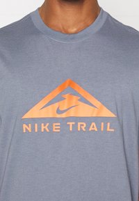 Acercamiento de una persona que lleva una camisa azul-gris con un logotipo triangular naranja y el texto "NIKE TRAIL" en el pecho.
