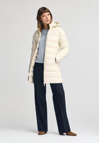 Cremefarvet pufferjakke med hætte, med en lynlås foran og to sidelommer. Båret over en lyseblå sweater og mørke bukser.