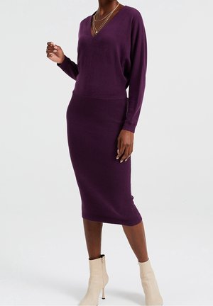 Lila, gerippter Strickkleid mit V-Ausschnitt und langen Ärmeln, mit einer taillierten Silhouette und strukturiertem Stoff. Kombiniert mit cremefarbenen Stiefeletten.