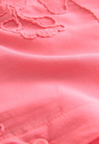 Koralo rožinės spalvos audinys su lygiu paviršiumi ir matomu gėlių siuvinėjimo raštu. Krašte yra sagtelė, atskleidžianti sudėtingą siuvimo darbą.